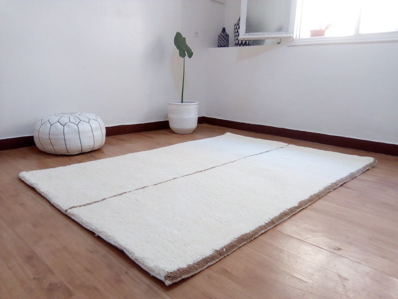 Puede incluir: Una alfombra de pelo blanco con una franja marr&oacute;n en el centro. La alfombra est&aacute; sobre un suelo de madera.