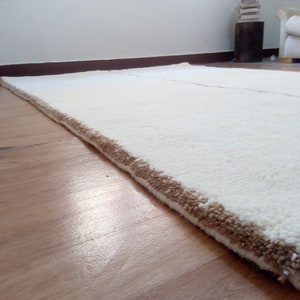 Puede incluir: Una alfombra blanca de pelo largo con un borde marr&oacute;n. La alfombra est&aacute; colocada sobre un suelo de madera.