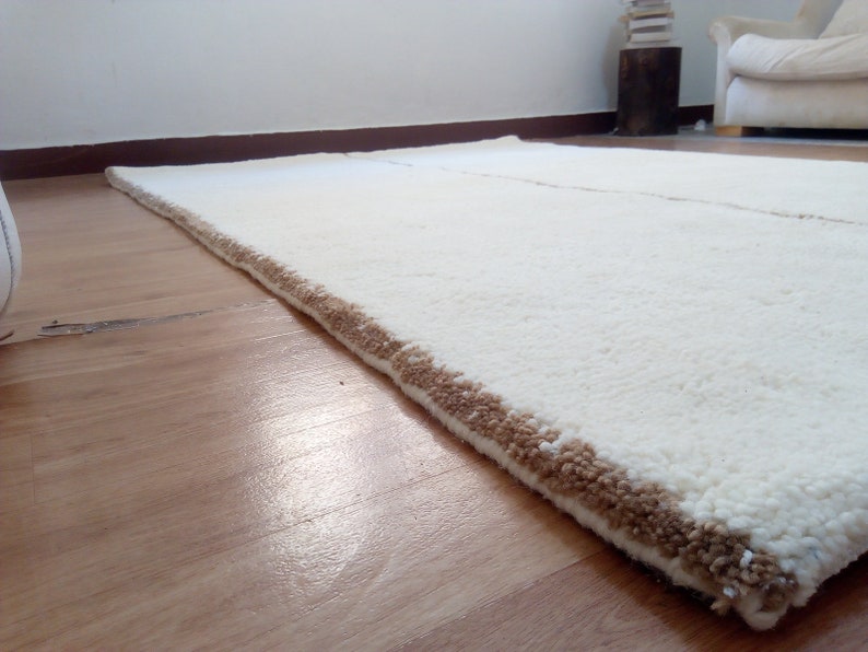 Puede incluir: Una alfombra blanca de pelo largo con un borde marr&oacute;n. La alfombra est&aacute; colocada sobre un suelo de madera.