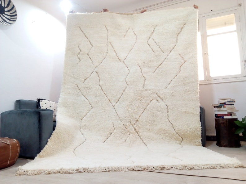 Puede incluir: Una alfombra marroqu&iacute; blanca con un patr&oacute;n geom&eacute;trico en un tono beige m&aacute;s claro. La alfombra tiene un borde de flecos y est&aacute; hecha de lana.