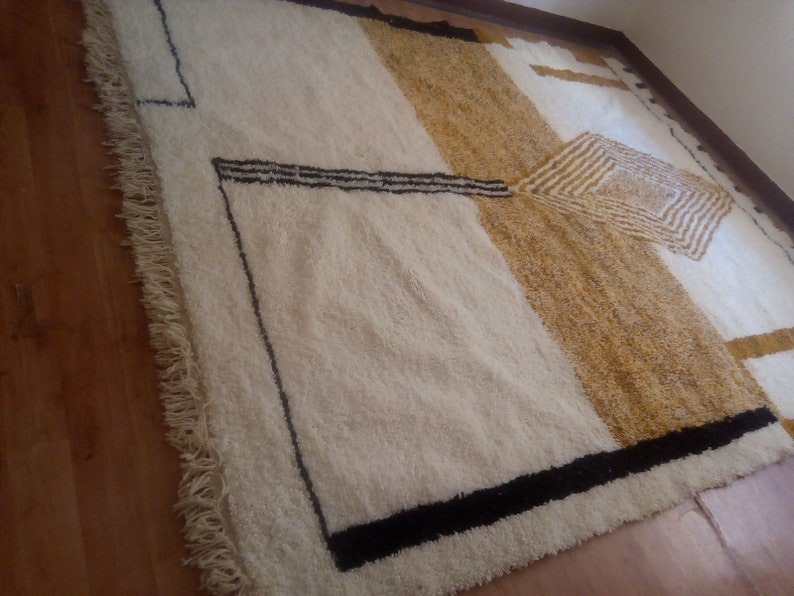 Puede incluir: Una alfombra blanca y beige con dise&ntilde;os geom&eacute;tricos negros. La alfombra tiene un borde de flecos.