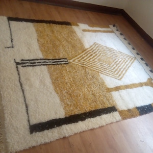 Puede incluir: Una alfombra blanca y dorada con un patr&oacute;n geom&eacute;trico. La alfombra tiene un borde de flecos y est&aacute; hecha de lana.
