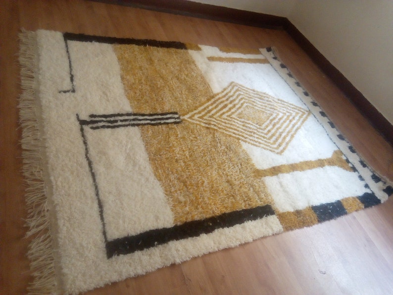 Puede incluir: Una alfombra blanca y dorada con un patr&oacute;n geom&eacute;trico. La alfombra tiene un borde de flecos y est&aacute; hecha de lana.