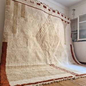 May include: Large, off-white rug with a geometric diamond pattern in the center. The rug has a thick, textured pile and is trimmed with a rust-colored border and tassels. The rug is approximately 8 feet by 10 feet.