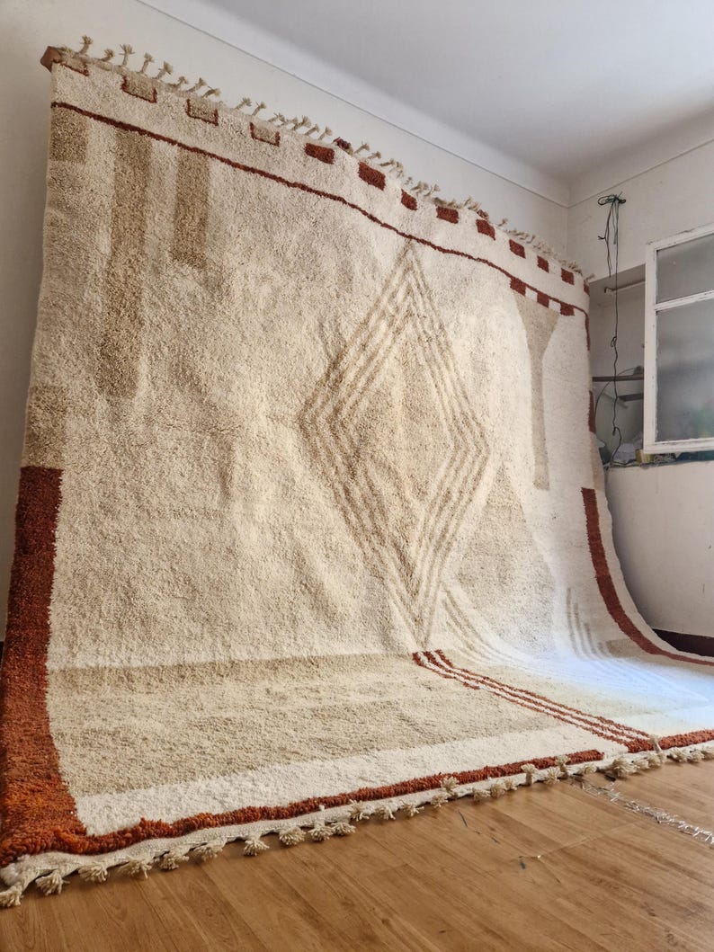 May include: Large, off-white rug with a geometric diamond pattern in the center. The rug has a thick, textured pile and is trimmed with a rust-colored border and tassels. The rug is approximately 8 feet by 10 feet.