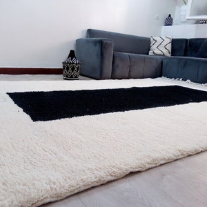 May include: A white and black Moroccan rug with a black rectangular design in the center. The rug has a fringe border and is made of wool.