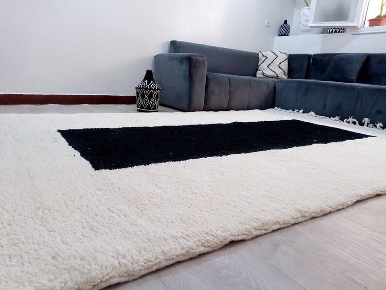 May include: A white and black Moroccan rug with a black rectangular design in the center. The rug has a fringe border and is made of wool.