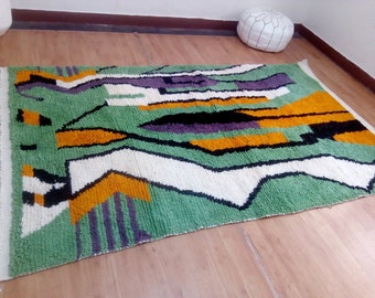 May include: Abstract geometric rug with a green background, white, black, orange, and purple accents. The rug has a fringed edge.