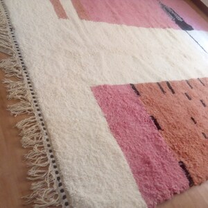 May include: A white rug with a geometric pattern in pink, orange, and black. The rug has a fringe border.