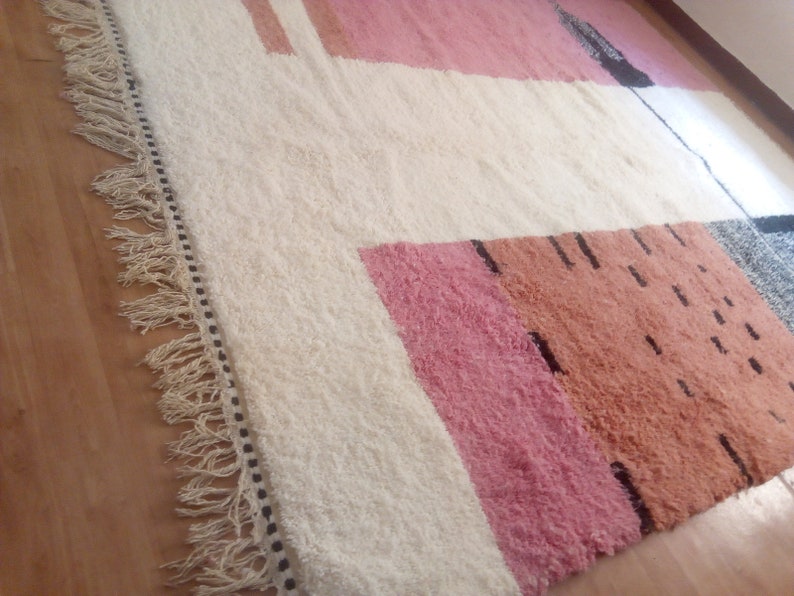 May include: A white rug with a geometric pattern in pink, orange, and black. The rug has a fringe border.