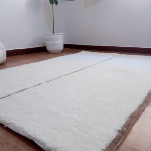 Puede incluir: Una alfombra de pelo blanco con un borde marr&oacute;n. La alfombra est&aacute; colocada sobre un suelo de madera.
