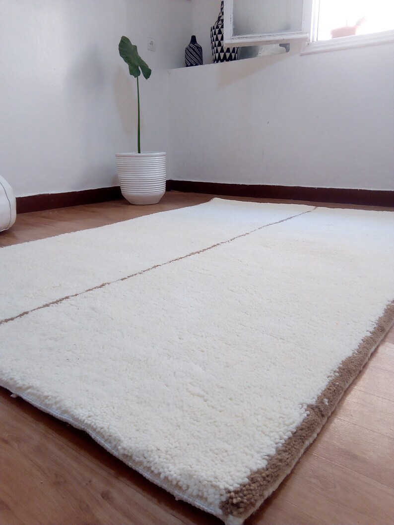Puede incluir: Una alfombra de pelo blanco con un borde marr&oacute;n. La alfombra est&aacute; colocada sobre un suelo de madera.