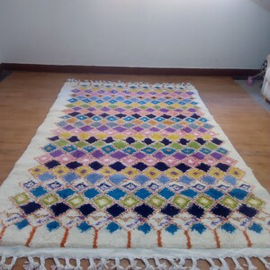 May include: A white rug with a colorful diamond pattern in shades of blue, green, pink, yellow, and purple. The rug has a fringe border.