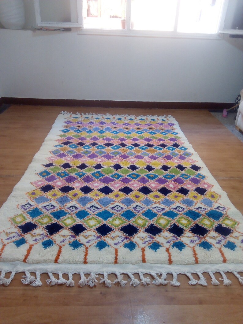 May include: A white rug with a colorful diamond pattern in shades of blue, green, pink, yellow, and purple. The rug has a fringe border.