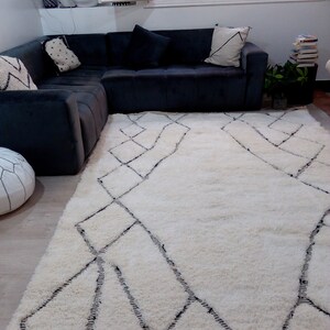 May include: A white shag rug with a black diamond pattern. The rug is in a living room with a black sectional sofa and a potted plant.