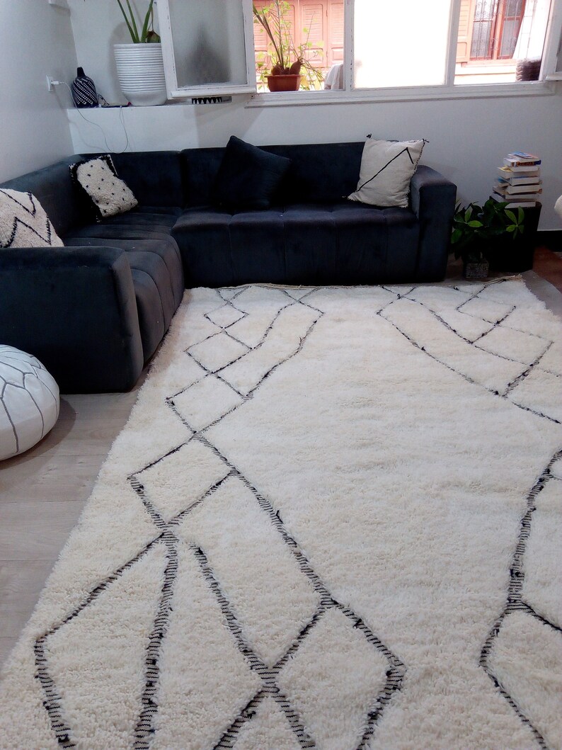 May include: A white shag rug with a black diamond pattern. The rug is in a living room with a black sectional sofa and a potted plant.