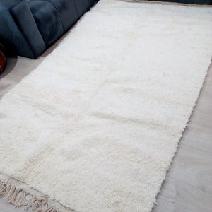 Pode incluir: Um tapete branco de pelo alto com uma borda de franjas. O tapete est&aacute; colocado sobre um piso de madeira clara.