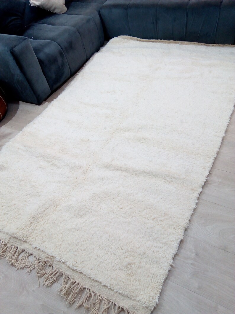 Pode incluir: Um tapete branco de pelo alto com uma borda de franjas. O tapete est&aacute; colocado sobre um piso de madeira clara.