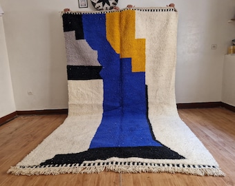 Pode incluir: Um tapete geométrico branco, azul, preto e amarelo com franjas nas bordas.