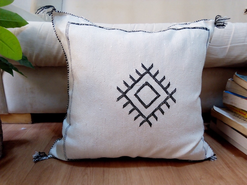 May include: White throw pillow with black embroidered geometric design. The pillow has black tassels on the corners.