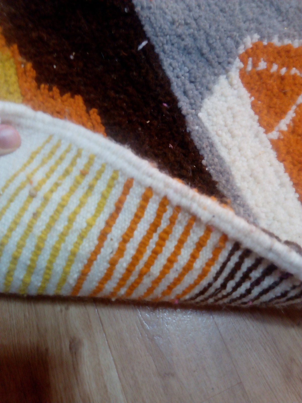 May include: A section of a colorful rug with a geometric pattern. The rug features a mix of colors including brown, orange, yellow, gray, and white. The edge of the rug is visible, showing a striped pattern in orange, yellow, and brown.