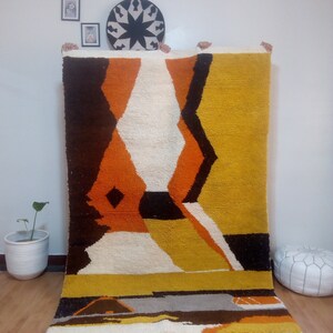 May include: A large, rectangular rug with a geometric design in shades of brown, orange, yellow, white, and black. The rug has a thick, textured pile and a fringe along the bottom edge. The design features diamond and angular shapes.