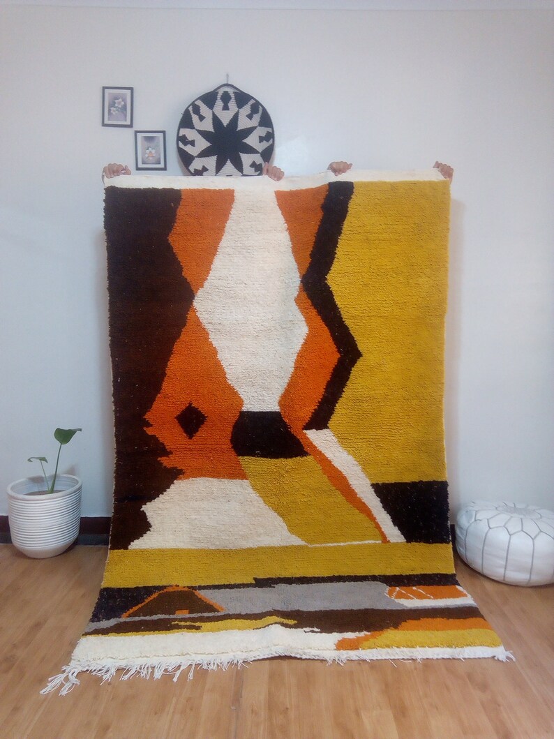 May include: A large, rectangular rug with a geometric design in shades of brown, orange, yellow, white, and black. The rug has a thick, textured pile and a fringe along the bottom edge. The design features diamond and angular shapes.