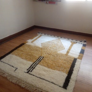 Puede incluir: Una alfombra geom&eacute;trica blanca y dorada con detalles negros y flecos en los bordes. La alfombra est&aacute; colocada sobre un suelo de madera.