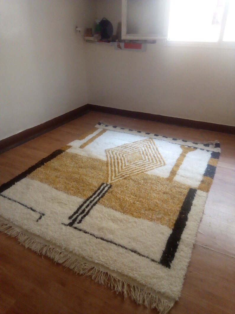 Puede incluir: Una alfombra geom&eacute;trica blanca y dorada con detalles negros y flecos en los bordes. La alfombra est&aacute; colocada sobre un suelo de madera.