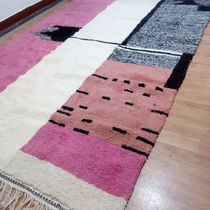 May include: A rectangular rug with a geometric pattern in shades of pink, white, and black. The rug has a fringe border and is made of wool.