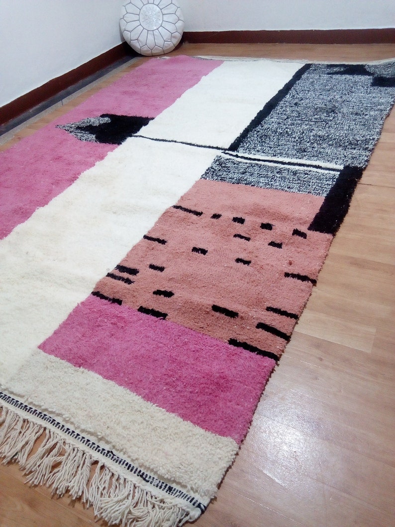 May include: A rectangular rug with a geometric pattern in shades of pink, white, and black. The rug has a fringe border and is made of wool.