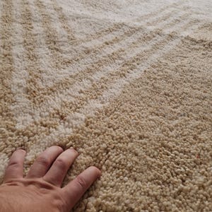 May include: A close-up of a beige rug with a textured, shaggy surface. The rug has a pattern of diagonal lines in a slightly darker shade of beige. The texture appears soft and plush, suggesting a comfortable floor covering.