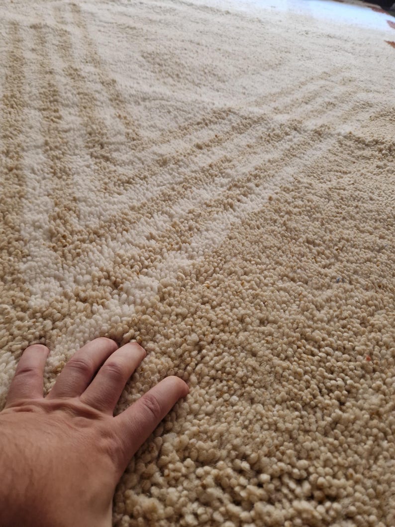 May include: A close-up of a beige rug with a textured, shaggy surface. The rug has a pattern of diagonal lines in a slightly darker shade of beige. The texture appears soft and plush, suggesting a comfortable floor covering.