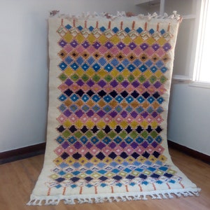 May include: A colorful Moroccan rug with a diamond pattern in shades of pink, blue, yellow, green, and black. The rug has a white background and fringe on the edges.