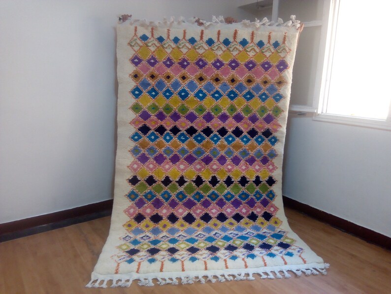 May include: A colorful Moroccan rug with a diamond pattern in shades of pink, blue, yellow, green, and black. The rug has a white background and fringe on the edges.