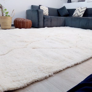 Puede incluir: Una alfombra blanca de pelo largo con un patr&oacute;n geom&eacute;trico. La alfombra est&aacute; en una sala de estar con un sof&aacute; seccional gris y un puf de cuero marr&oacute;n.