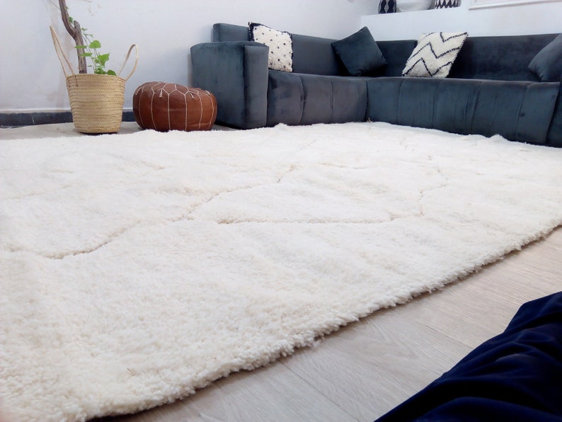 Puede incluir: Una alfombra blanca de pelo largo con un patr&oacute;n geom&eacute;trico. La alfombra est&aacute; en una sala de estar con un sof&aacute; seccional gris y un puf de cuero marr&oacute;n.