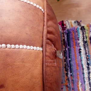 May include: A brown leather pouf with white stitching and a zipper on the side. The pouf is sitting on a colorful woven rug.
