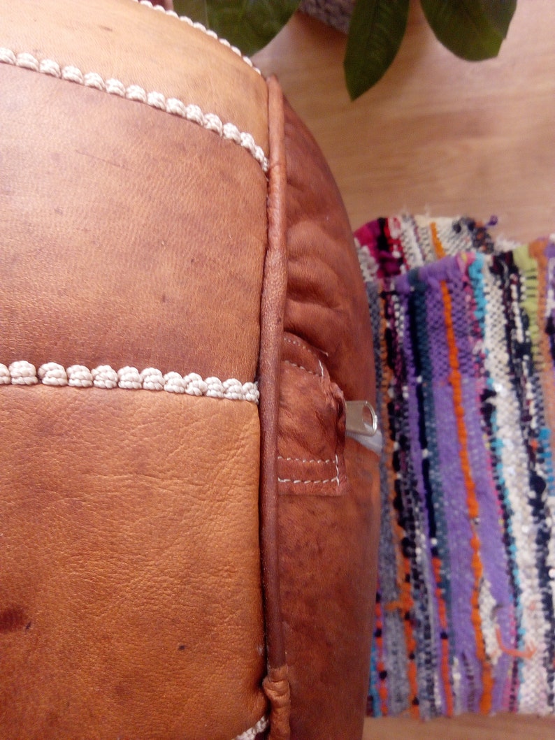 May include: A brown leather pouf with white stitching and a zipper on the side. The pouf is sitting on a colorful woven rug.