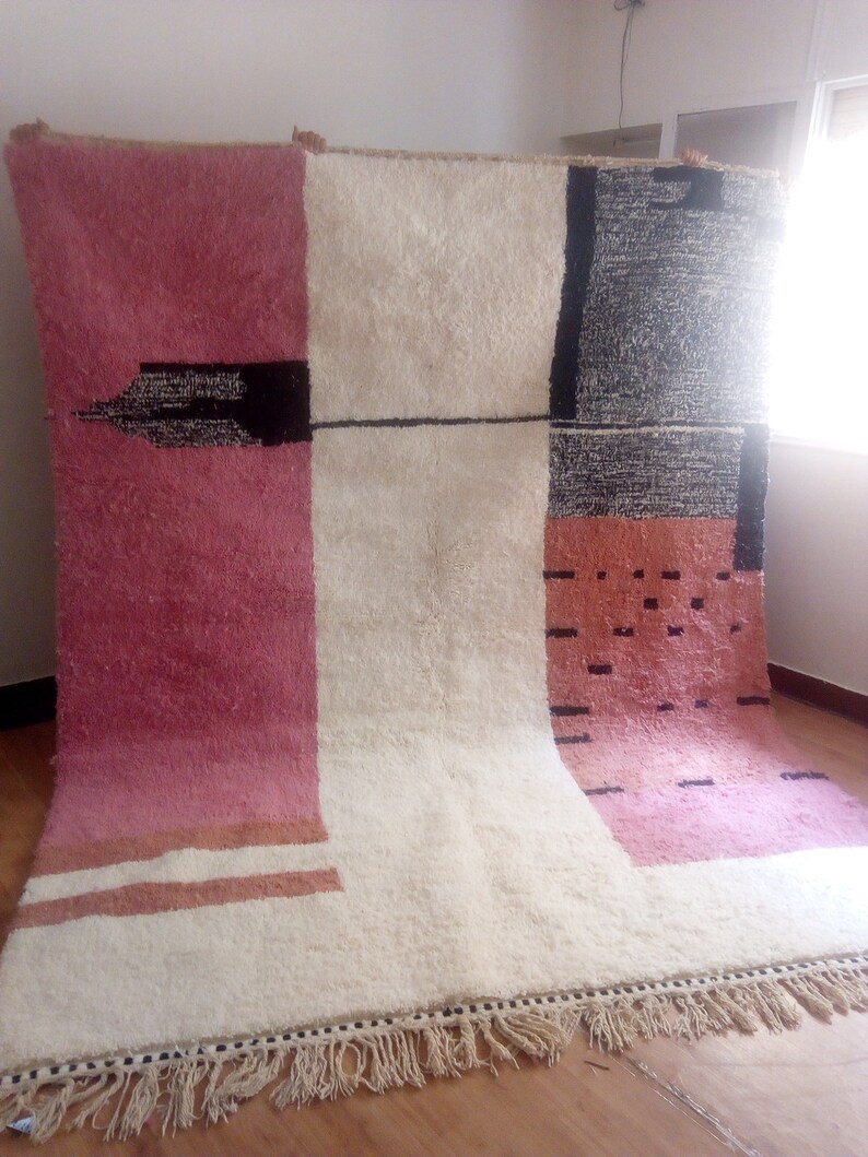 May include: A white, pink, and black geometric patterned rug with a fringe border. The rug has a modern design with a mix of colors and textures.