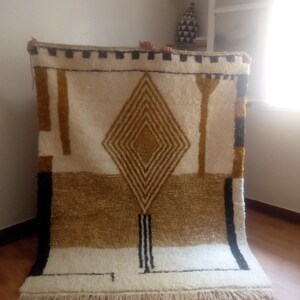 Puede incluir: Una alfombra geom&eacute;trica blanca, crema y dorada con detalles negros. La alfombra tiene un patr&oacute;n de diamante en el centro y un borde de flecos.
