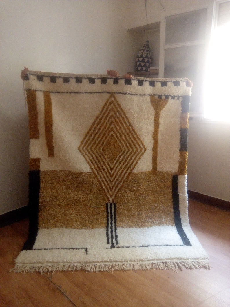 Puede incluir: Una alfombra geom&eacute;trica blanca, crema y dorada con detalles negros. La alfombra tiene un patr&oacute;n de diamante en el centro y un borde de flecos.