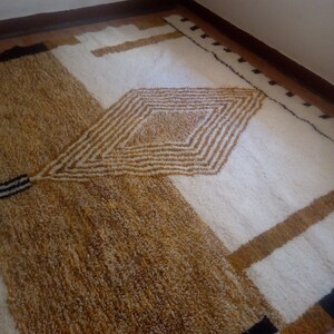 Puede incluir: Una alfombra de pelo blanco y marr&oacute;n con un patr&oacute;n geom&eacute;trico. La alfombra tiene una forma de diamante en el centro con rayas y un borde negro.