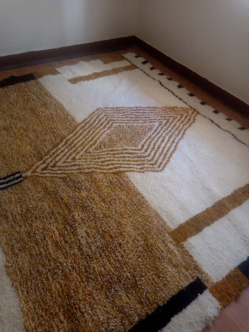 Puede incluir: Una alfombra de pelo blanco y marr&oacute;n con un patr&oacute;n geom&eacute;trico. La alfombra tiene una forma de diamante en el centro con rayas y un borde negro.
