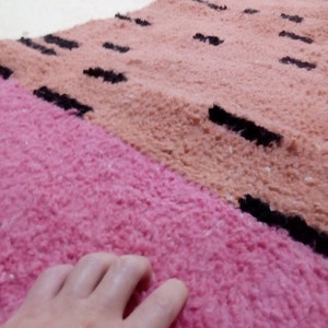 May include: Close-up of a pink and peach colored rug with black stripes. A hand is resting on the pink section of the rug.