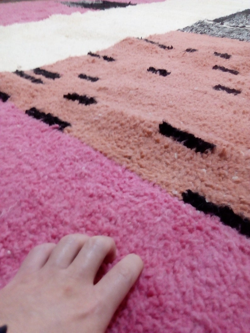 May include: Close-up of a pink and peach colored rug with black stripes. A hand is resting on the pink section of the rug.
