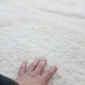 Pode incluir: Um close-up de um tapete branco e felpudo com uma m&atilde;o descansando na superf&iacute;cie. O tapete tem uma textura macia e felpuda.