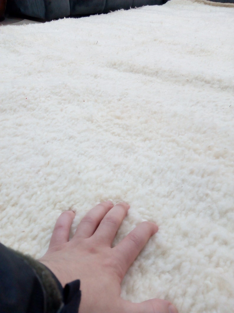 Pode incluir: Um close-up de um tapete branco e felpudo com uma m&atilde;o descansando na superf&iacute;cie. O tapete tem uma textura macia e felpuda.