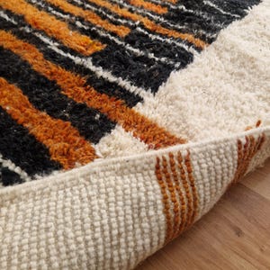 May include: Close-up of the edge of a white, orange, and black patterned rug. The rug is folded over, showing the white backing and the orange and black stripes of the fringe.