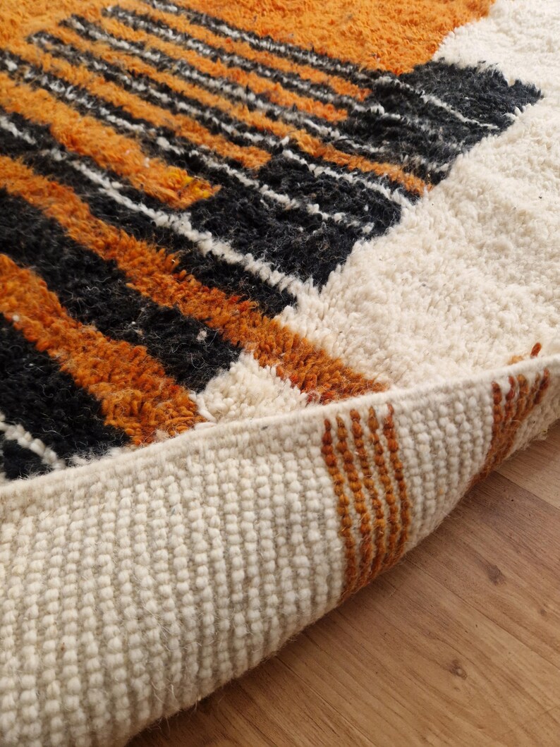 May include: Close-up of the edge of a white, orange, and black patterned rug. The rug is folded over, showing the white backing and the orange and black stripes of the fringe.